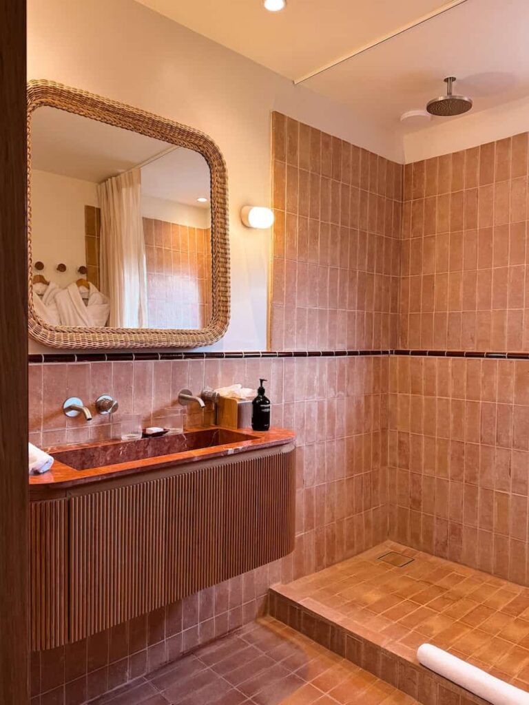 A bathroom with terracotta tiles, a frameless mirror with a woven frame, and a walk in shower with a rain shower head, at capelongue hotel. 