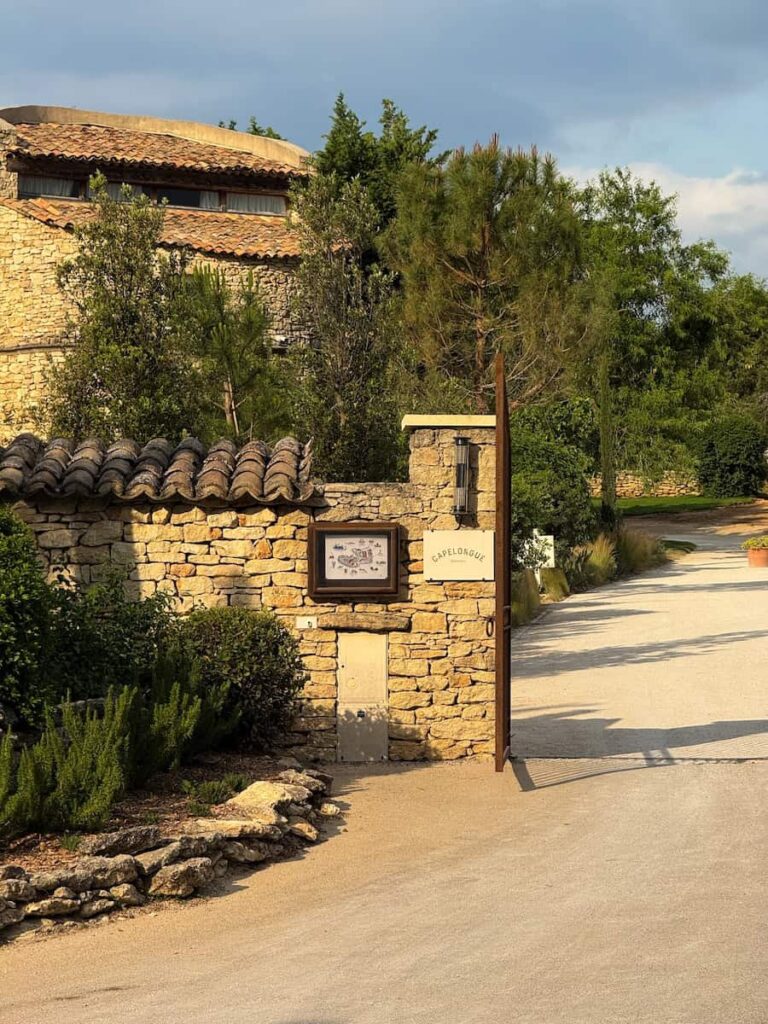 A stone entrance gate with a Capelongue sign and surrounding greenery under soft late afternoon light.