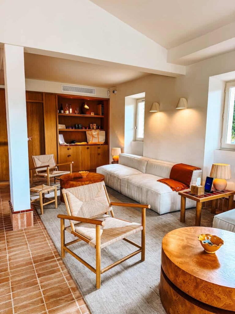 Lobby of Capelongue hotel with natural wood furniture, woven chairs, a cream sofa, and shelves displaying decor in warm earthy tones.