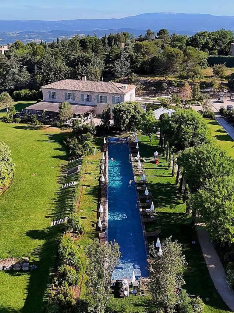 A long outdoor swimming pool surrounded by green lawns, trees, and lounge chairs with a stone house and distant hills in the background.