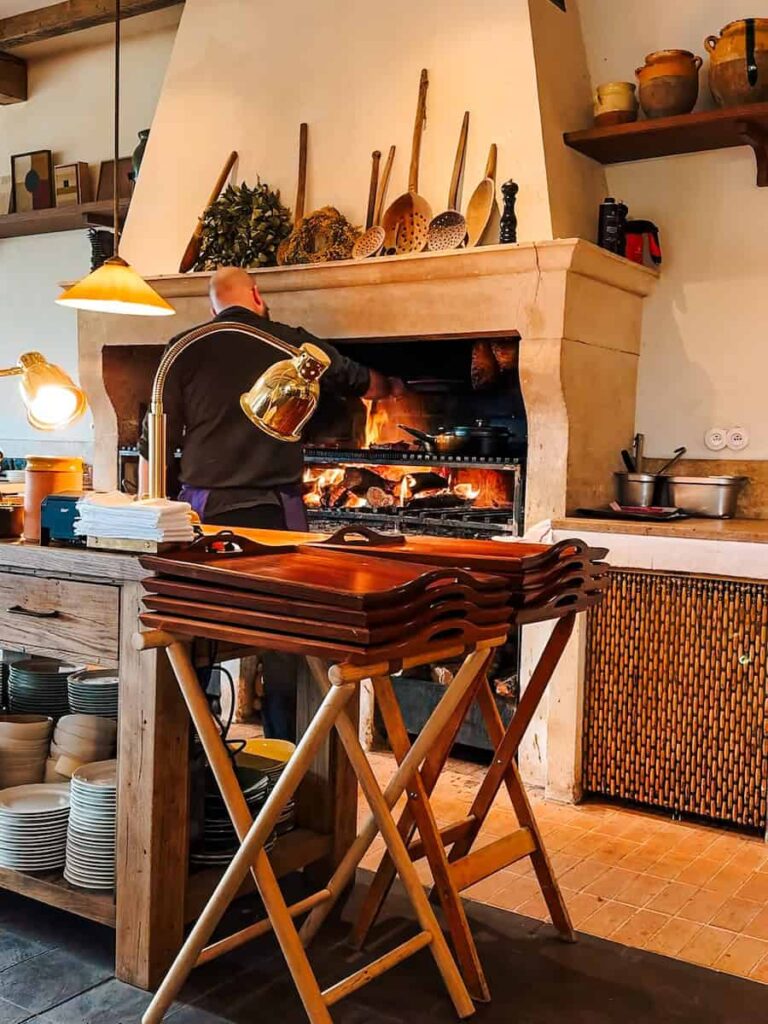 A chef cooking over an open fire grill inside a rustic kitchen with wooden trays and shelves of pottery.