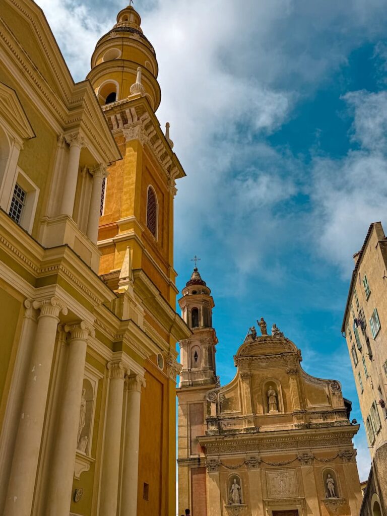 View of the Basilica of Saint Michel in Menton from below, showing its ornate architecture and tall bell tower against a blue sky.