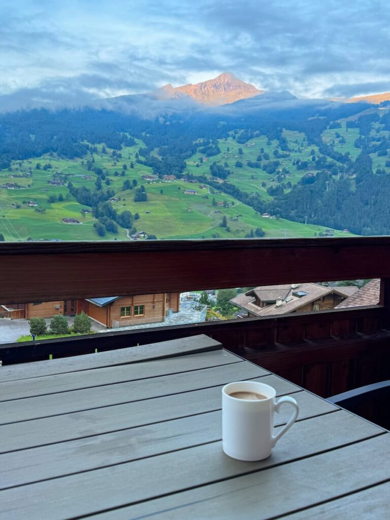 Cup of coffee on a chalet balcony in Grindelwald overlooking green alpine meadows and sunlit mountain peaks.