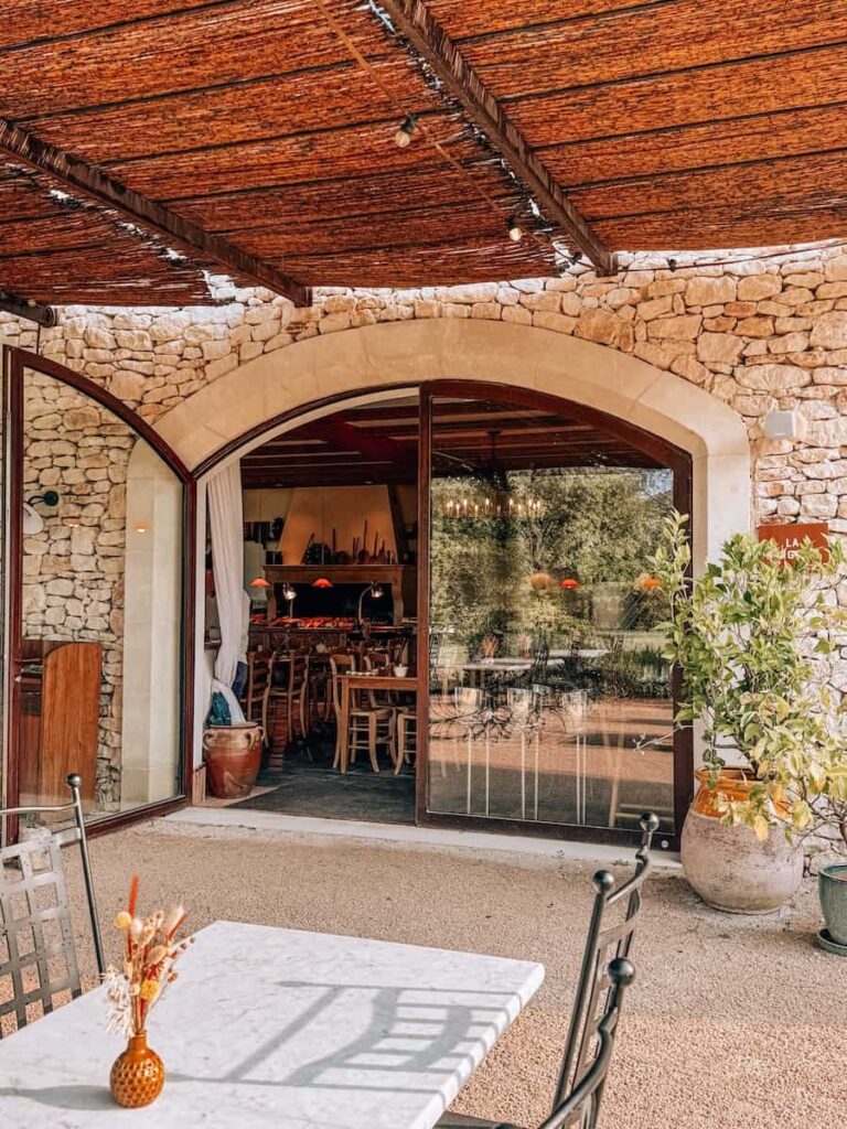 A shaded outdoor terrace with a stone wall and arched glass doorway leading into a cozy dining space.