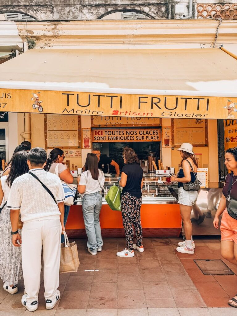 People lining up at the Tutti Frutti ice cream stand in Menton, with a cream-colored awning and handwritten signs.