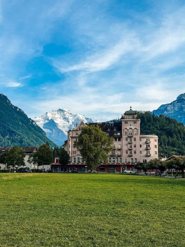 Large open meadow in Interlaken with a grand historic hotel and snow capped mountains towering in the background.
