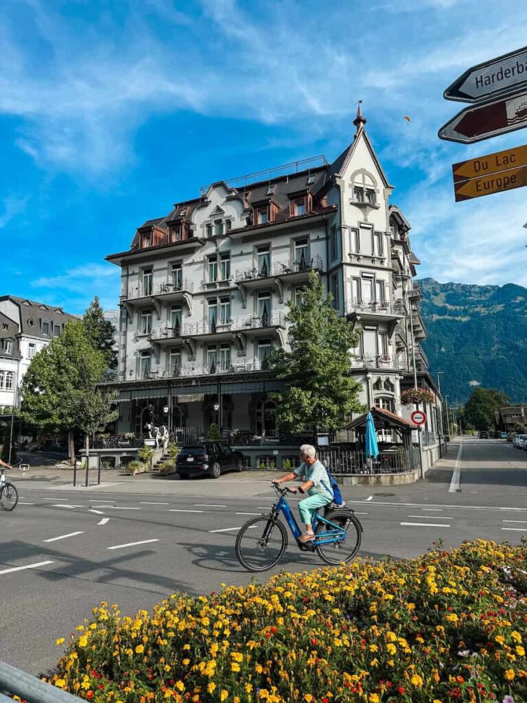 Charming historic building in Interlaken with cyclists passing by and bright yellow flowers in the foreground against a backdrop of alpine mountains.