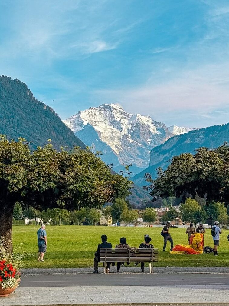 People relaxing in a park in Interlaken with paragliders landing and a stunning view of snow capped mountains in the distance.