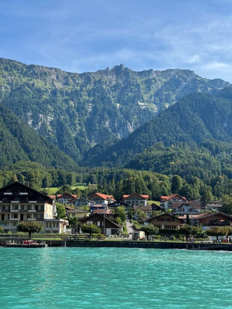 Turquoise waters of Lake Brienz with charming Swiss lakeside houses at the base of tall green mountains.