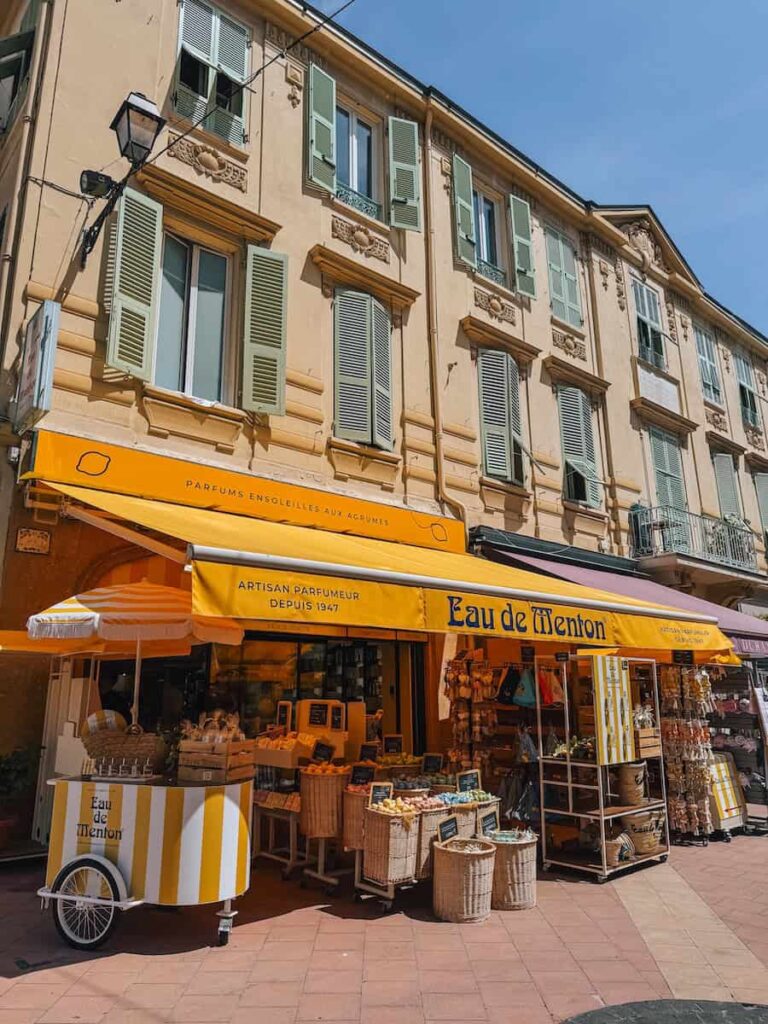 The Eau de Menton perfumery storefront with bright yellow awnings, citrus displays, and pastel shuttered windows above.