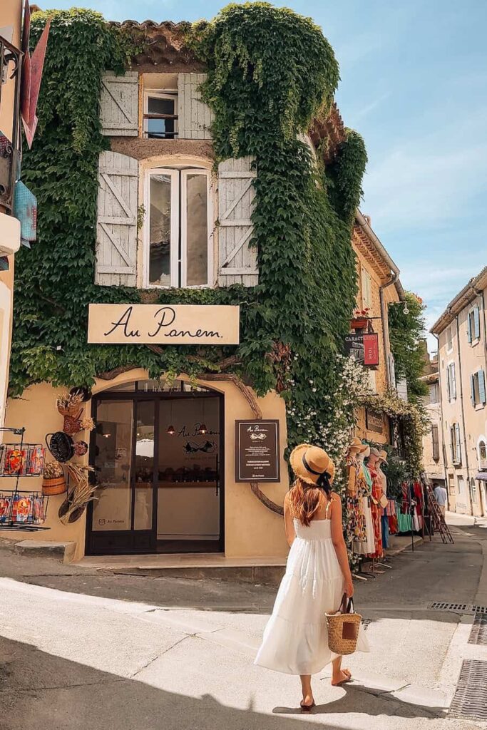 A woman in a white summer dress walking toward a shop covered in ivy along a charming village street.