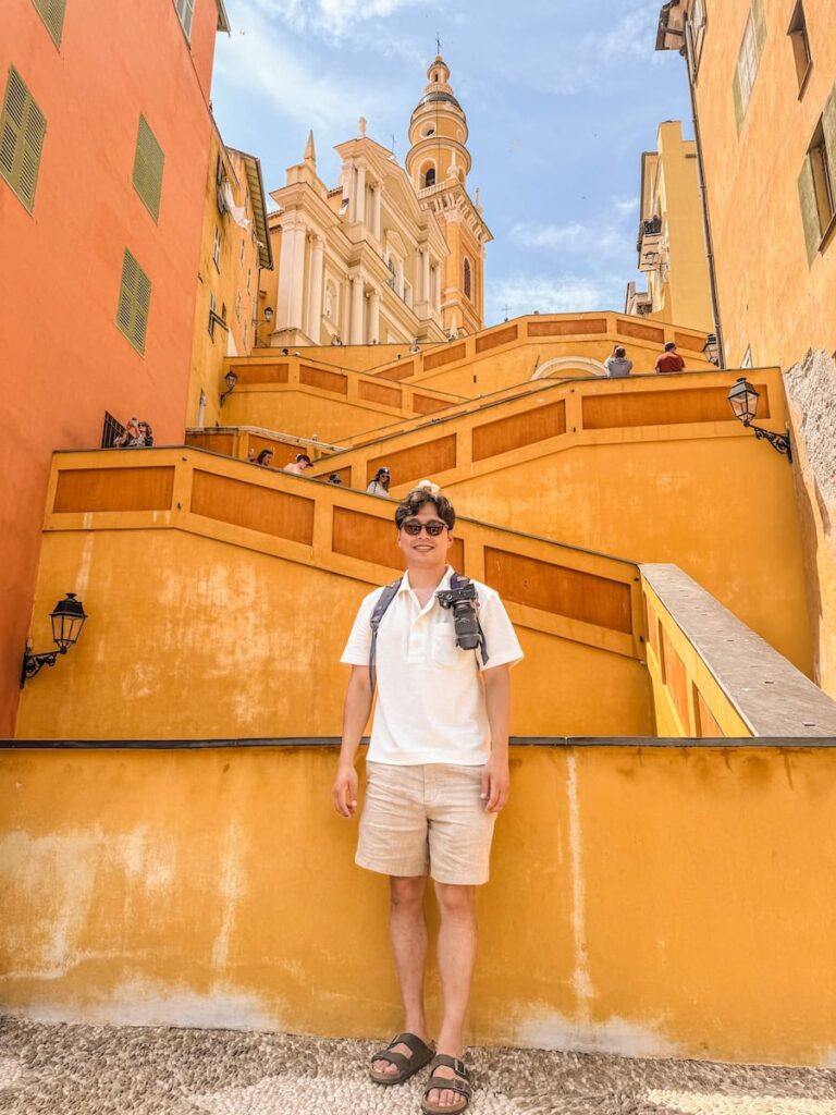 A man wearing sunglasses, a white shirt, and beige shorts standing in front of the yellow zigzag staircase and basilica in Menton.