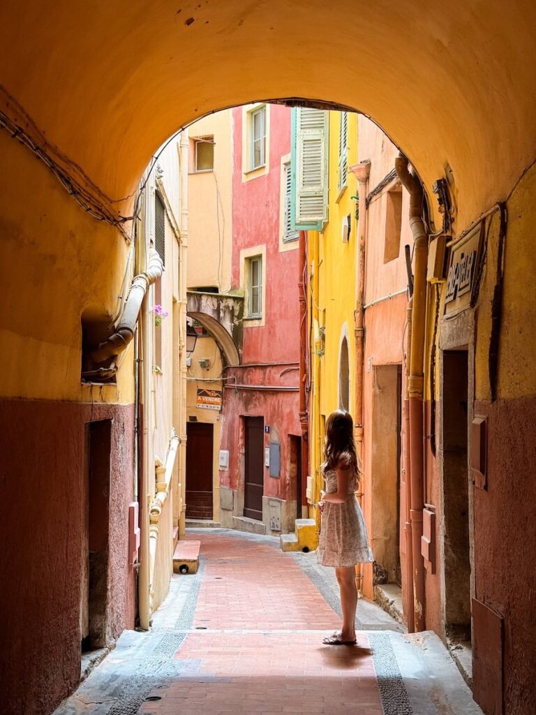 A woman in a light summer dress standing in a narrow alleyway in Menton France, surrounded by warm pastel buildings in shades of yellow and coral.