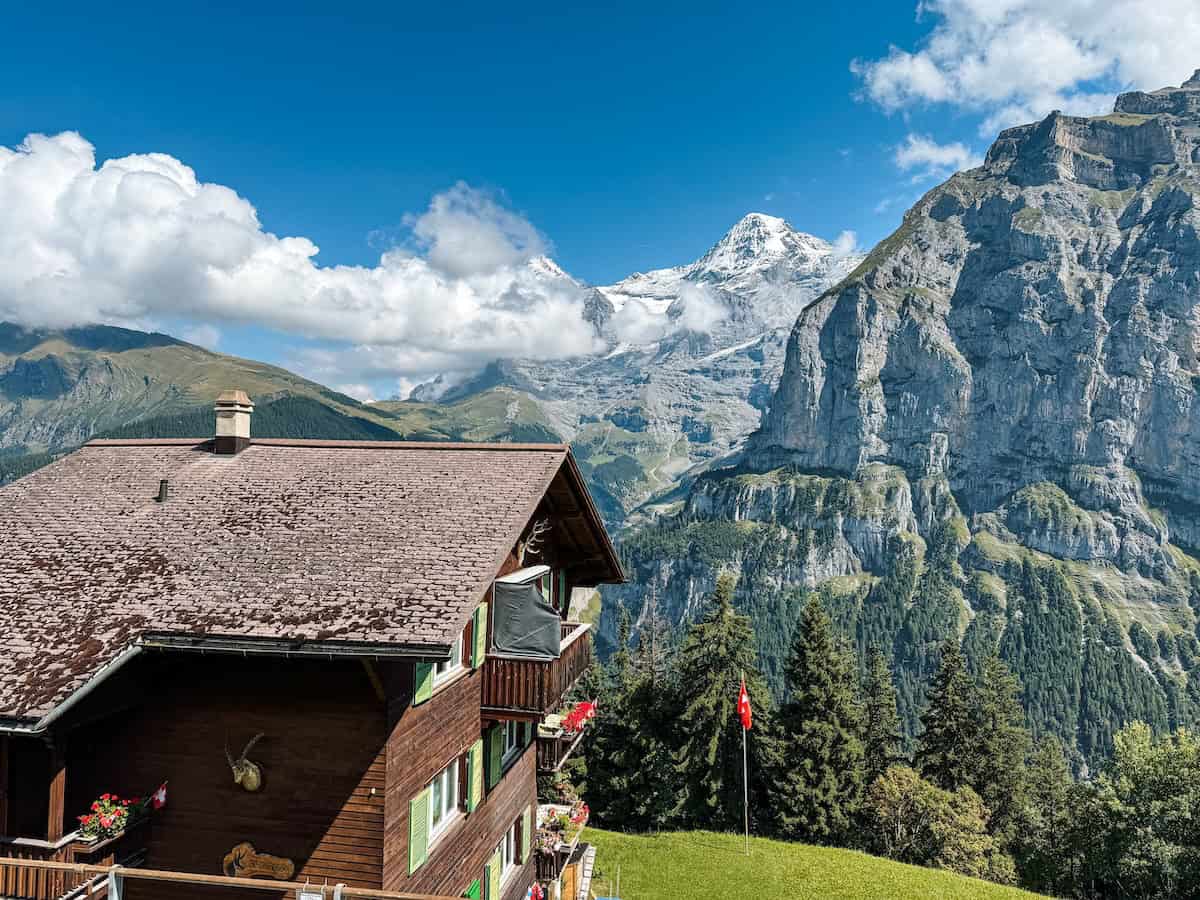 Charming wooden chalet in Mürren with bright flower boxes and sweeping views of towering alpine cliffs and snowy mountain peaks under a clear blue sky.