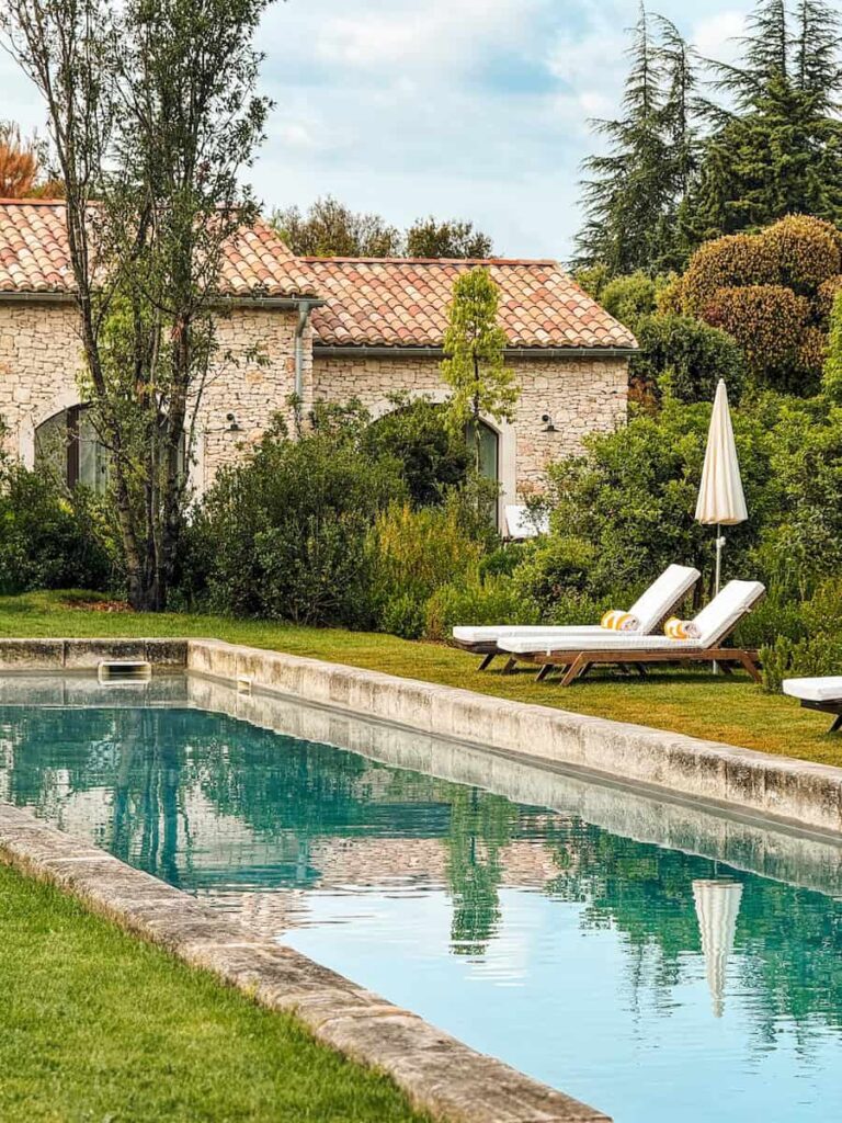 A long stone edged swimming pool surrounded by garden greenery, with white loungers and umbrellas beside a rustic stone building.