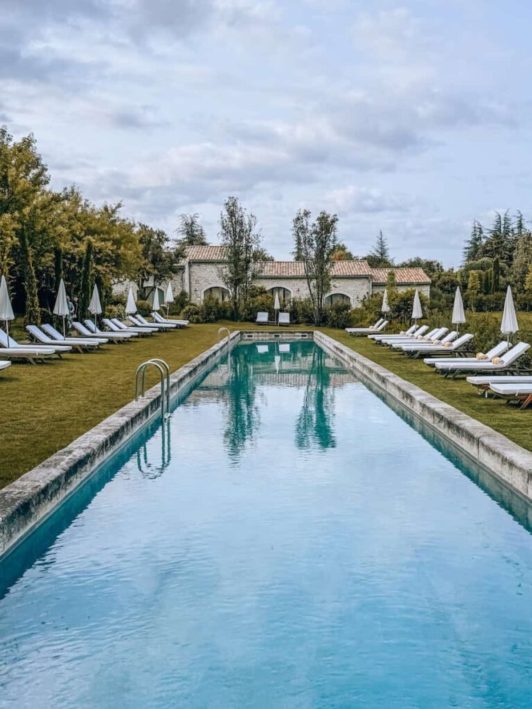A long rectangular swimming pool lined with white loungers and umbrellas, with stone buildings in the background.