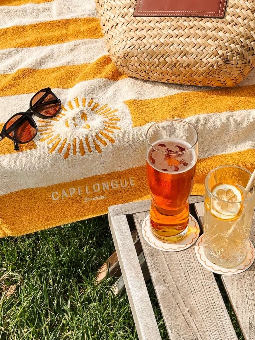 A striped yellow and white Capelongue pool towel with sunglasses, a straw bag, and two cold drinks set on a wooden side table in the grass.