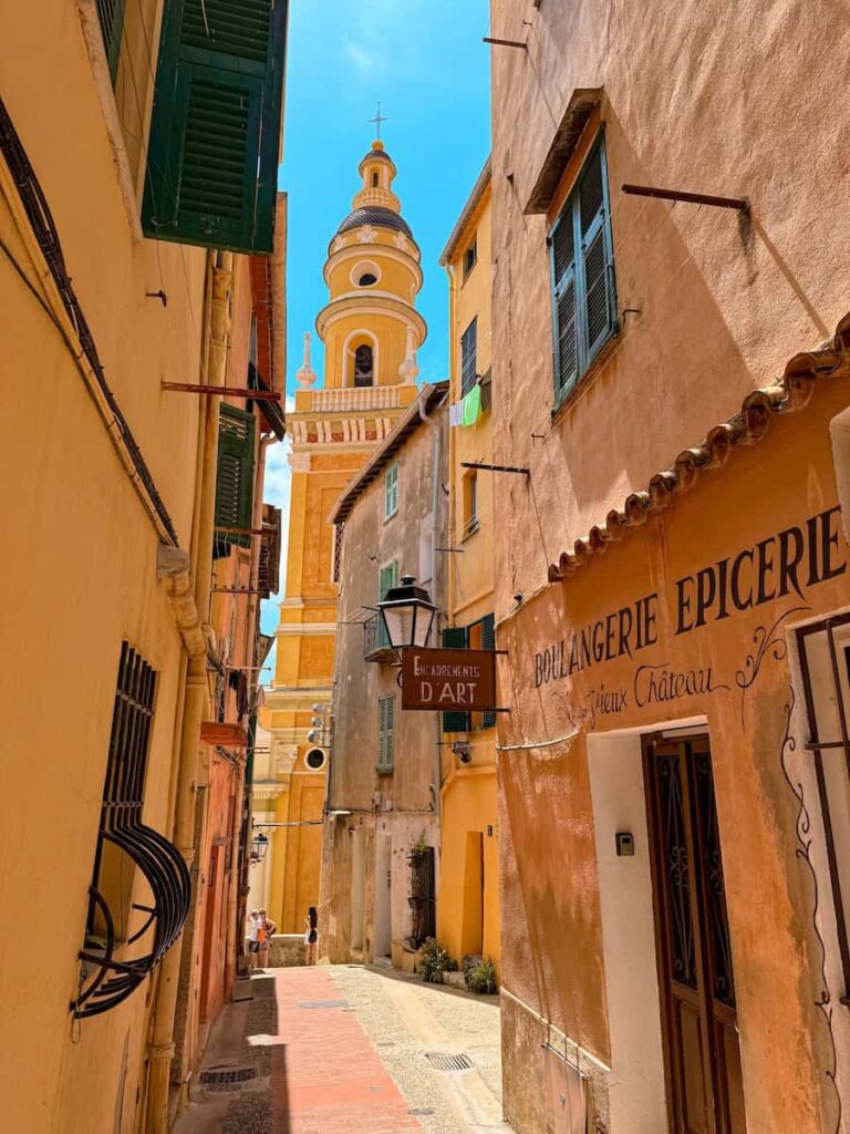 A narrow pastel street in Menton with warm yellow and peach buildings, leading toward the bell tower of the Basilica of Saint Michel under a bright blue sky.