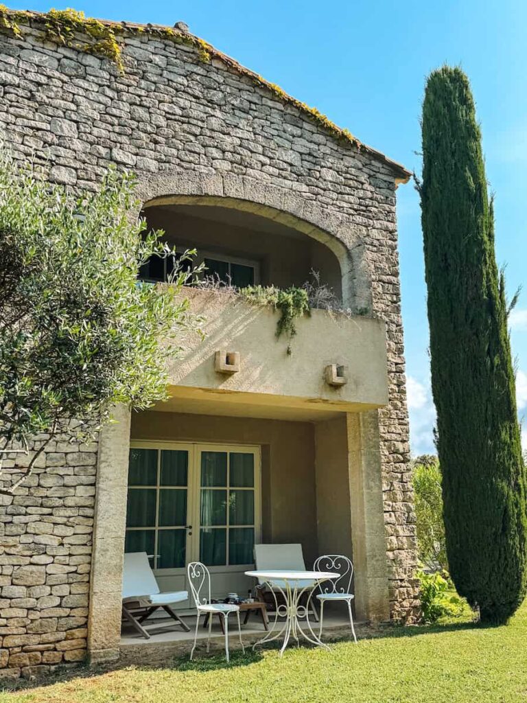 A stone farmhouse with a shaded terrace and white outdoor chairs overlooking a grassy garden.