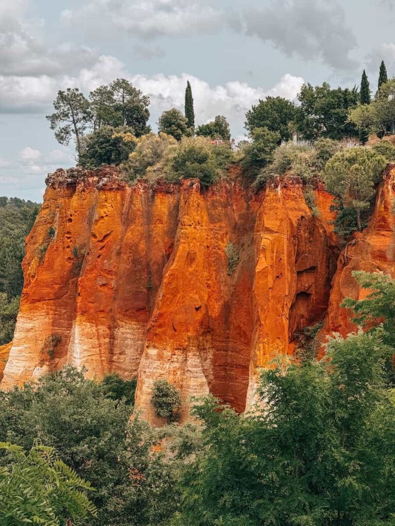 The dramatic ochre cliffs of Roussillon rising above lush green trees under a cloudy sky.