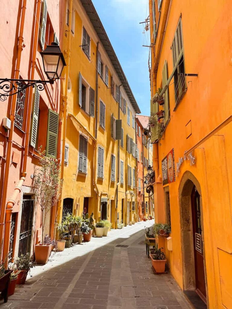 Narrow street in Menton lined with bright orange and yellow buildings, potted plants, and pastel shutters under a sunny sky.