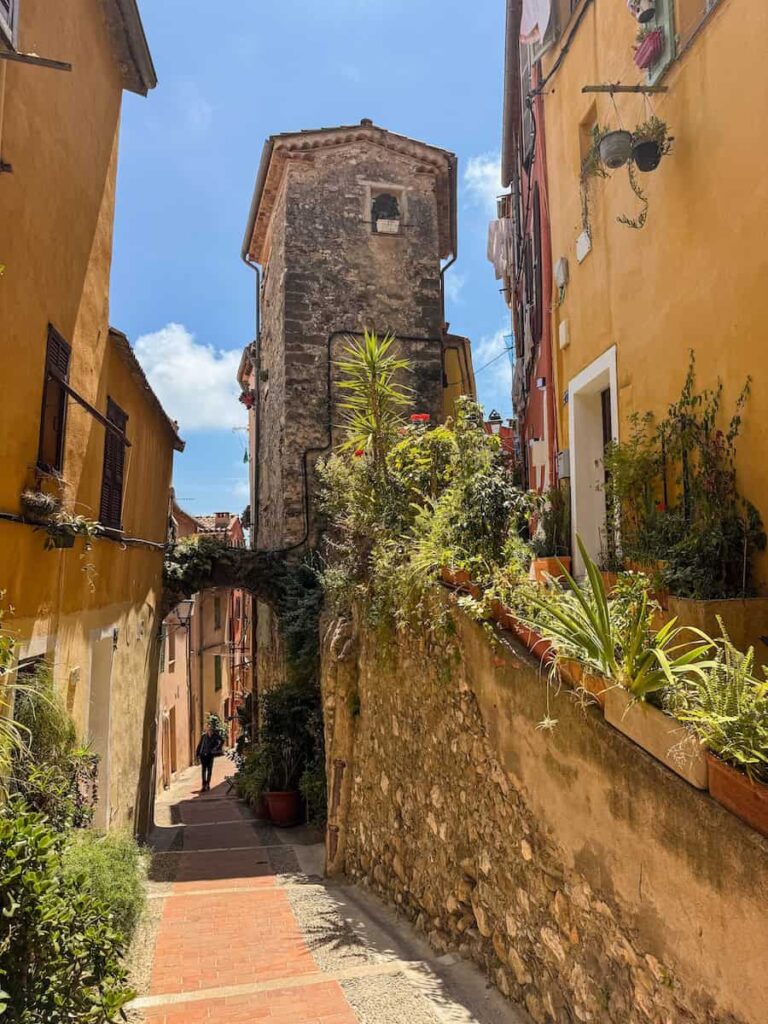 A quiet medieval pathway in Menton with rustic stone walls, potted plants, and an old stone archway leading into a shaded alley.