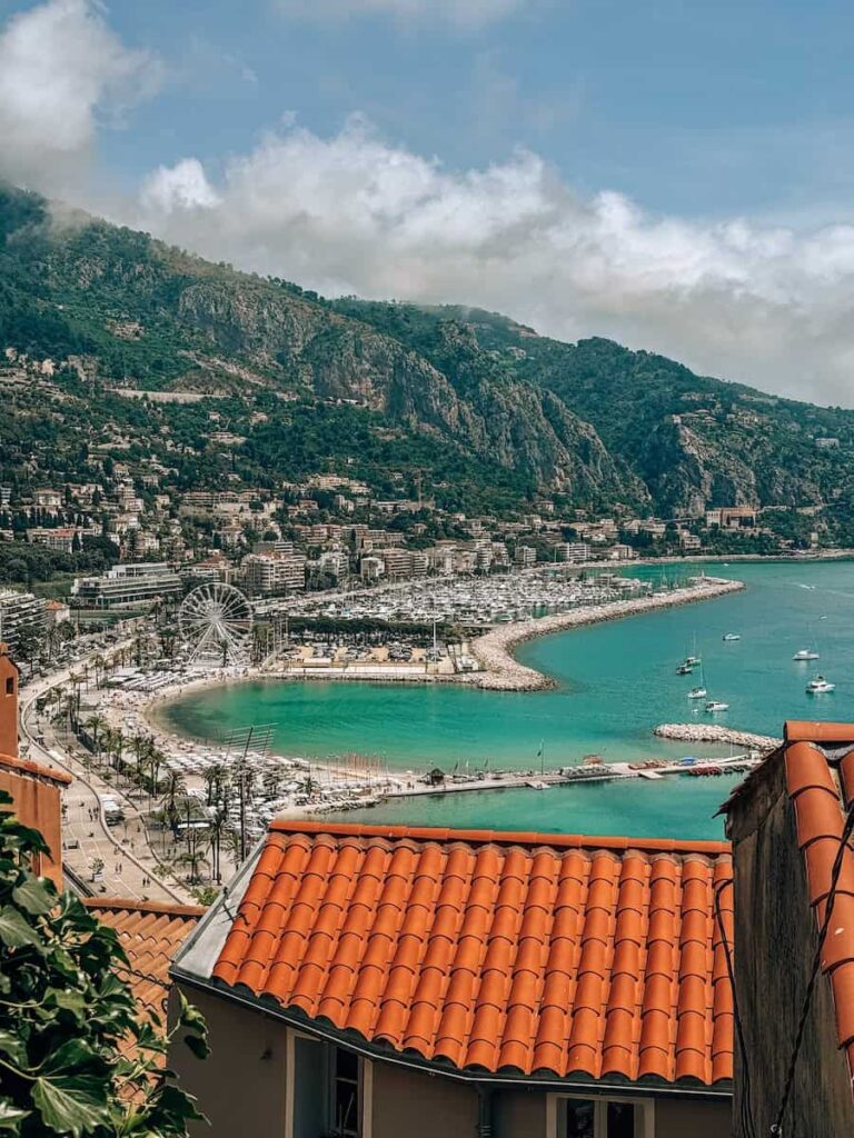 View of Menton’s coastline and marina from above, showing turquoise water, a curved beach, terracotta rooftops, and mountains in the distance.