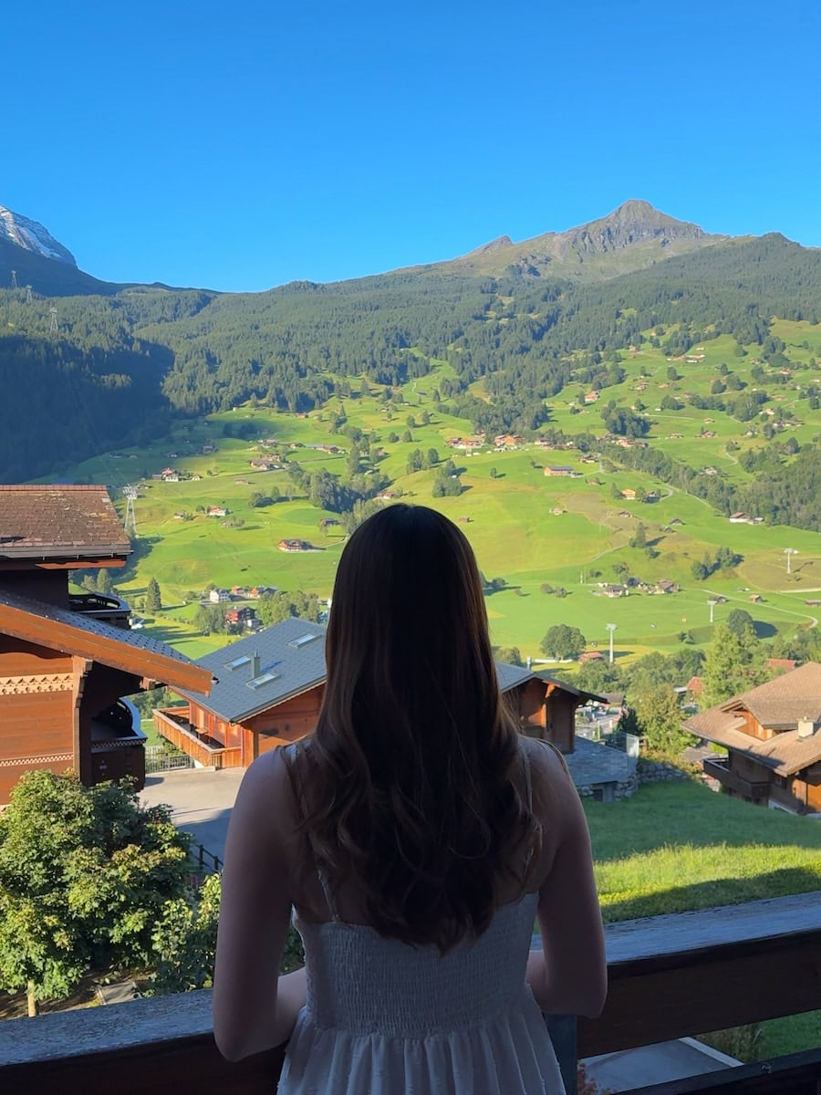 Author in a white dress standing on a balcony in Grindelwald, overlooking bright green alpine hills and wooden chalets under a clear blue sky.