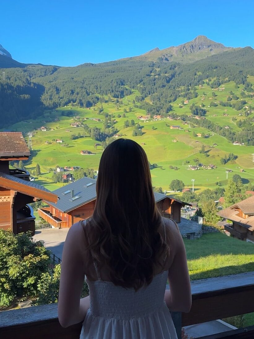Woman standing on a balcony and gazing out at the bright green valley and mountain slopes of Grindelwald on a clear morning.