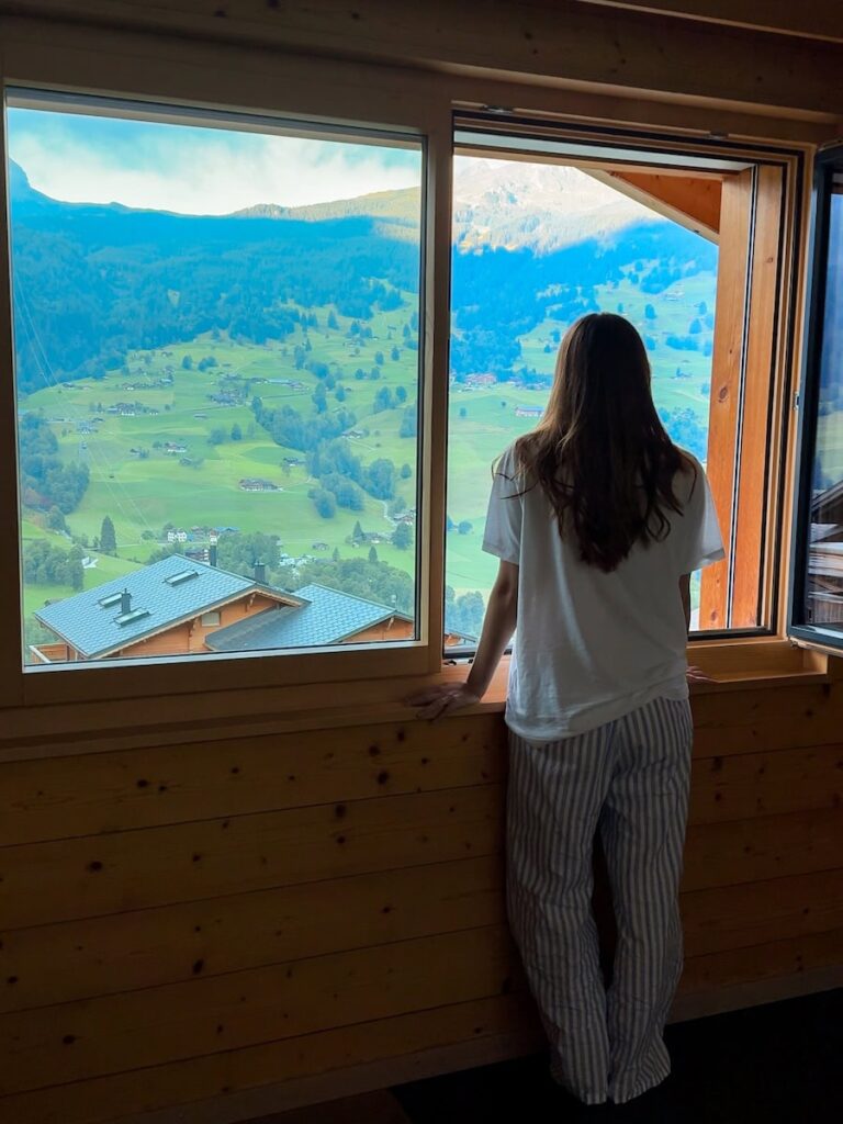 Woman in pajamas standing at a chalet window and overlooking the green alpine valley and mountain slopes of Grindelwald.