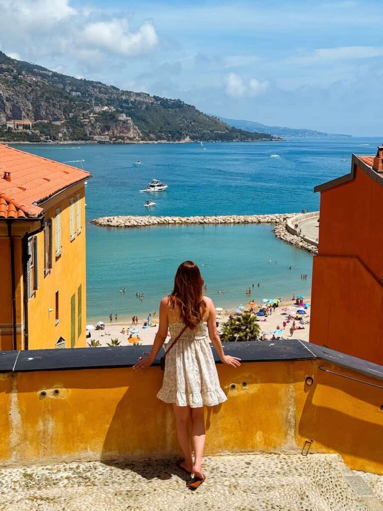 The author in a white summer dress standing at a lookout point in Menton, overlooking the turquoise water and beach below.