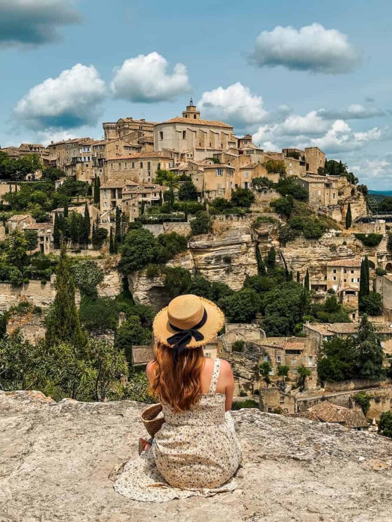 A woman in a straw hat and floral dress sitting on a rock ledge overlooking the hilltop village of Gordes.