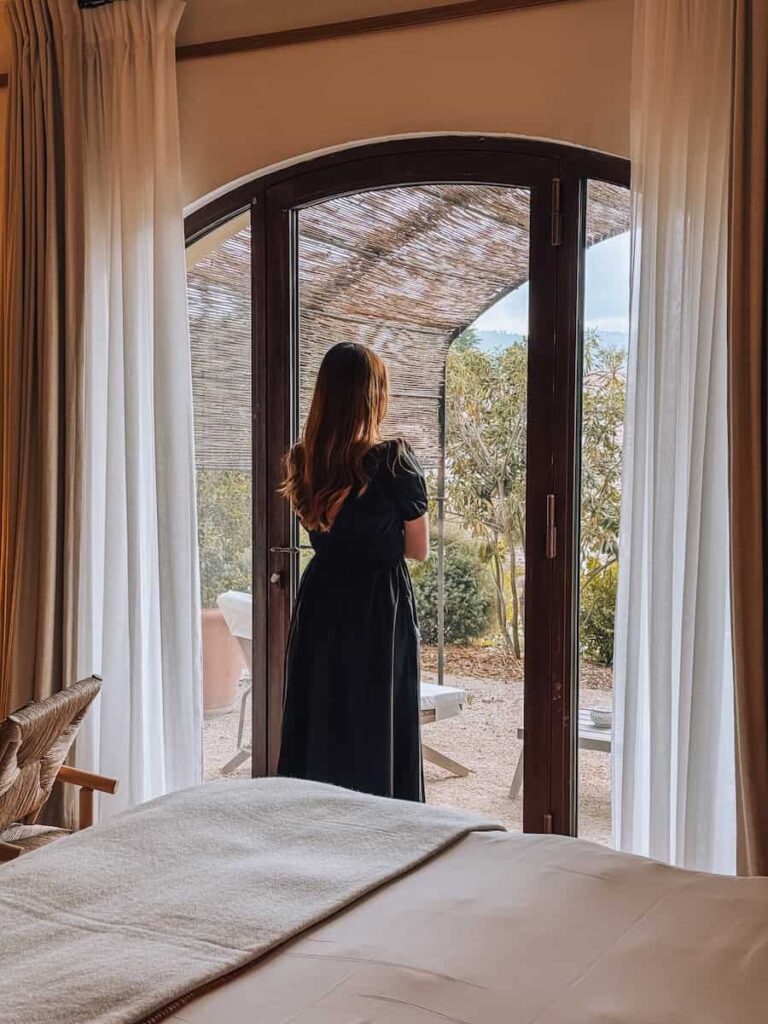 A woman in a black dress standing by glass doors, looking out toward a garden patio from the bedroom of Capelongue hotel. 