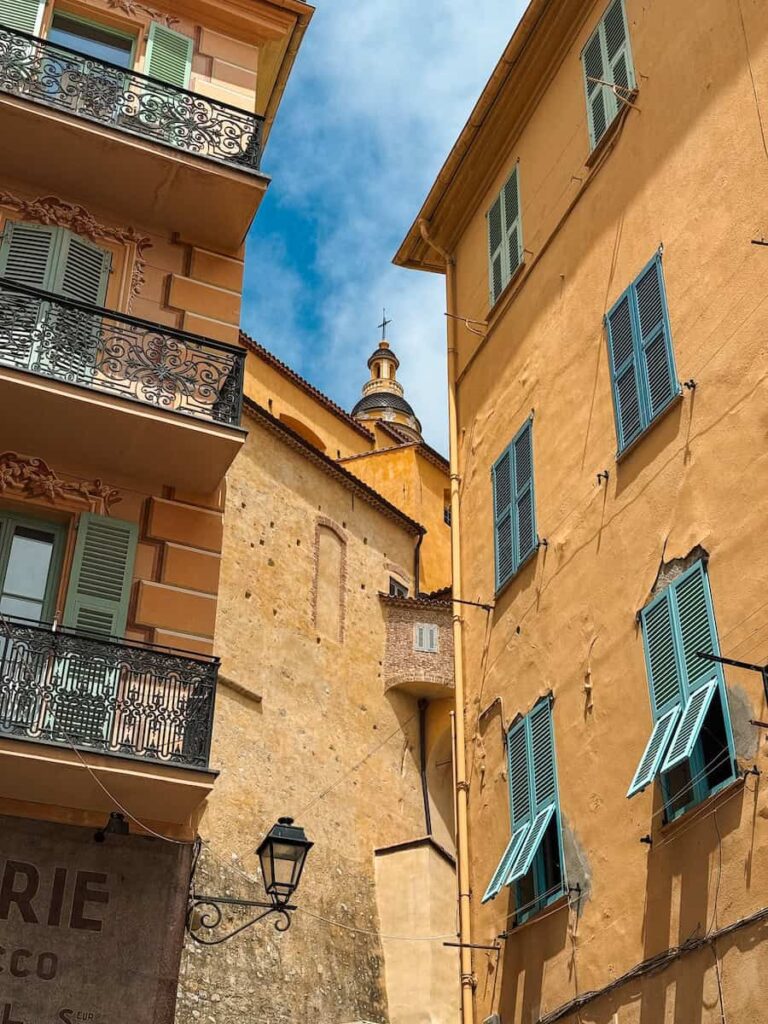 View of pastel buildings in Menton France with open green shutters and the basilica bell tower rising between them.