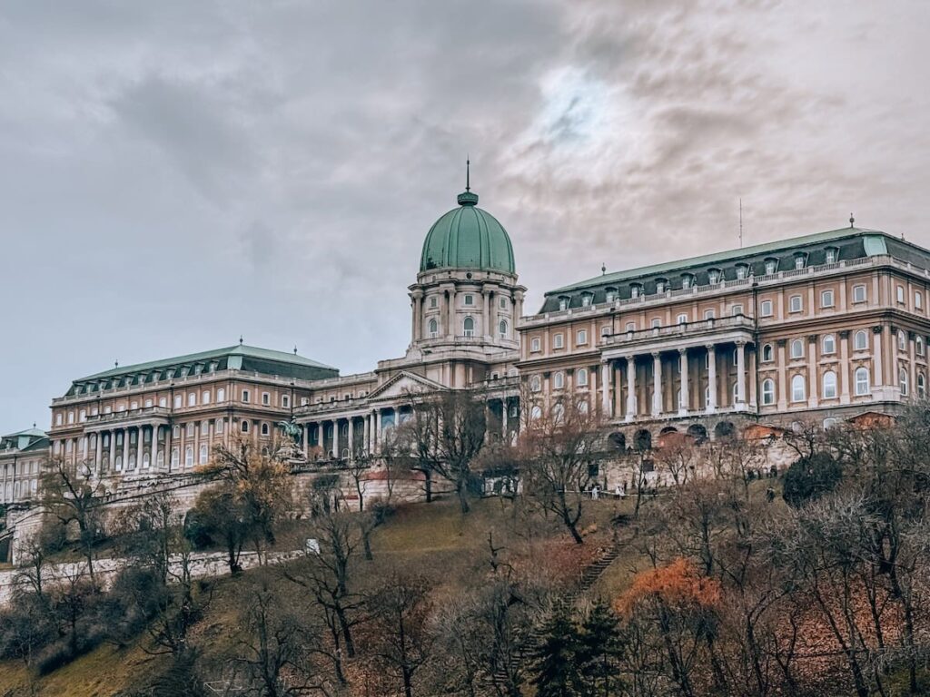 View of Buda Castle in Budapest perched on Castle Hill, featuring the grand historic palace with its green domed roof, classical facade, and terraced slopes below under an overcast sky.