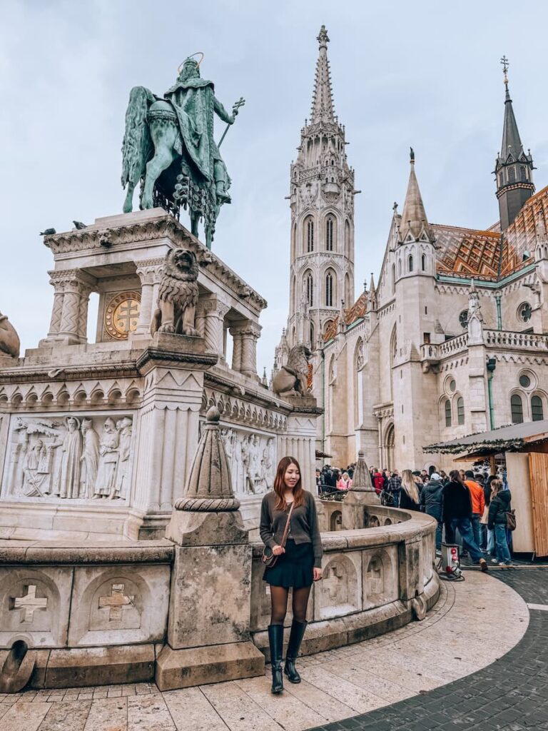 A woman standing near the statue of King Saint Stephen in the Buda Castle District with Matthias Church rising behind her and visitors gathered around the historic square.