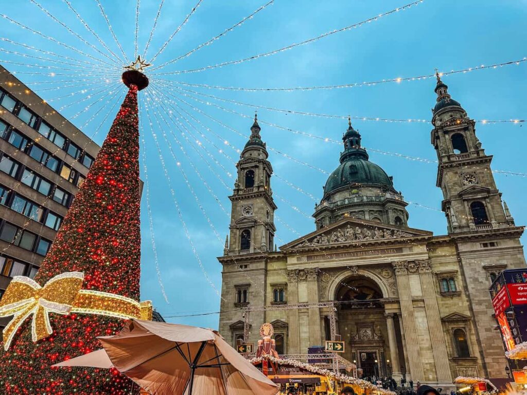 Christmas market at St Stephen’s Basilica in Budapest with a towering decorated Christmas tree, strings of festive lights overhead, and the illuminated basilica facade rising behind the stalls at dusk.
