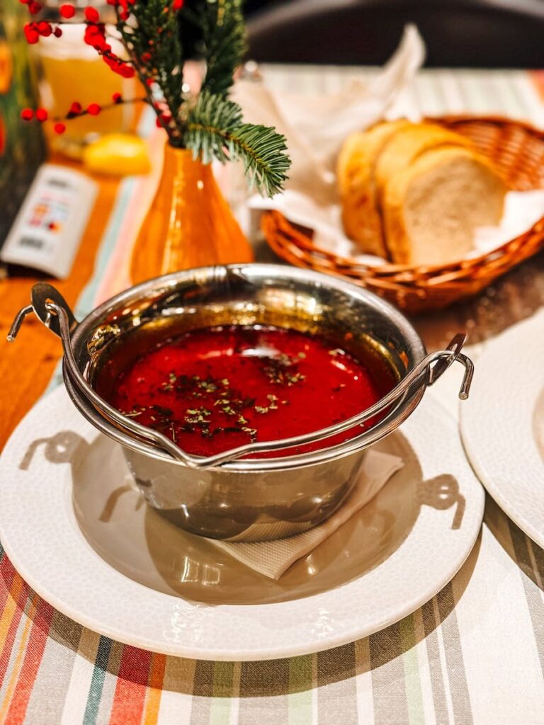 Traditional Hungarian goulash served in a small metal pot on a white plate, filled with rich red broth and herbs, with sliced bread in a basket and festive table decor in the background.