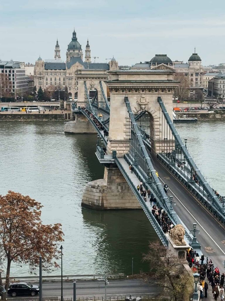 A view of the Chain Bridge stretching across the Danube River with pedestrians crossing and the Pest skyline visible in the background on an overcast day.