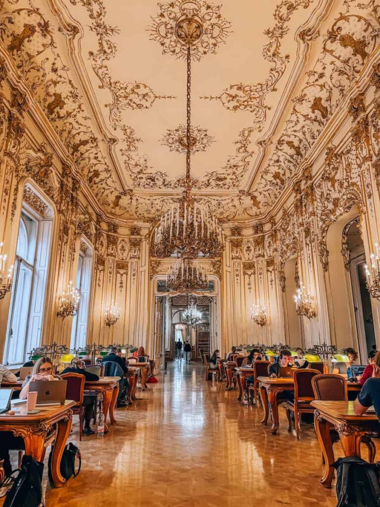 Grand reading room inside the Ervin Szabó Library in Budapest with ornate Baroque ceilings, crystal chandeliers, polished parquet floors, and long wooden desks where visitors are quietly studying.