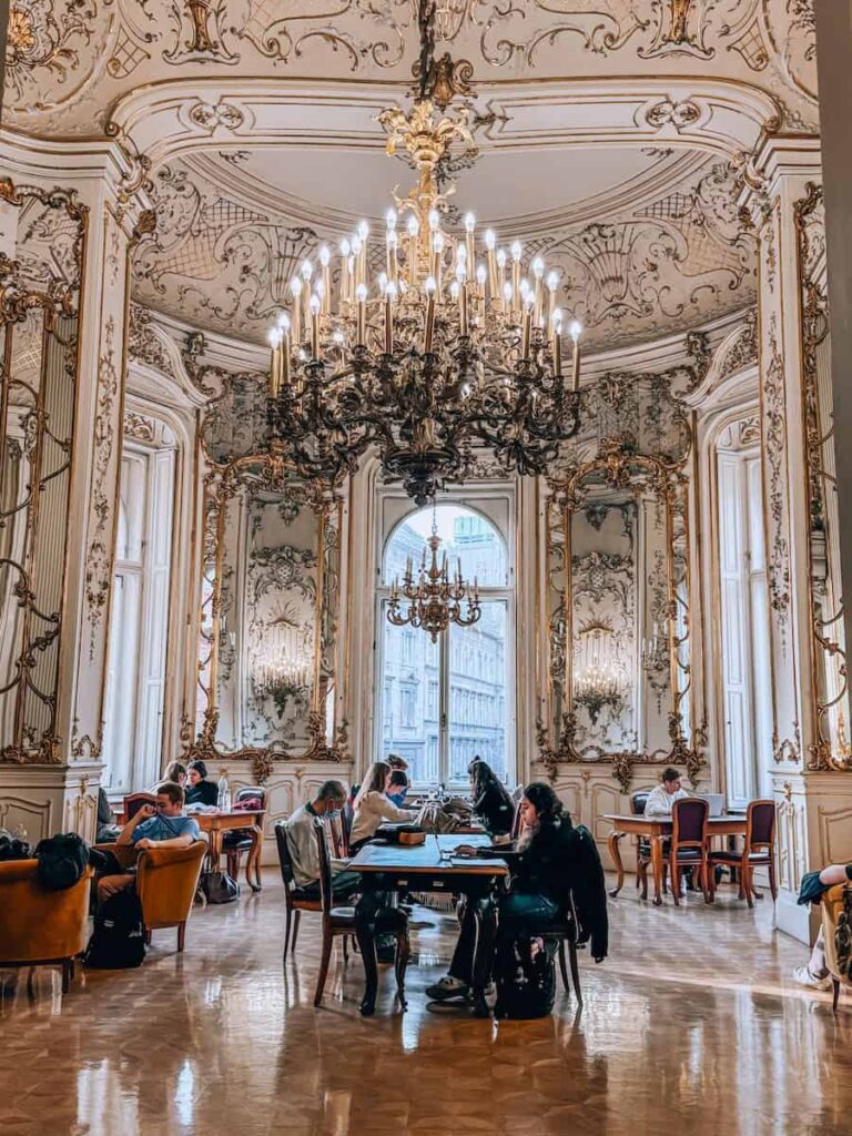 Elegant salon at the Ervin Szabó Library featuring an elaborate chandelier, gilded wall details, tall windows, and people seated at tables reading and working in a historic palace setting.