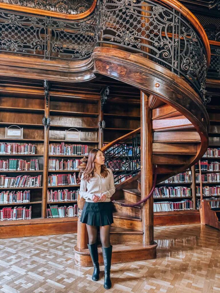 Woman standing beneath a sweeping wooden spiral staircase inside the Ervin Szabó Library, surrounded by floor to ceiling bookshelves and warm wood paneling in a classic library interior.