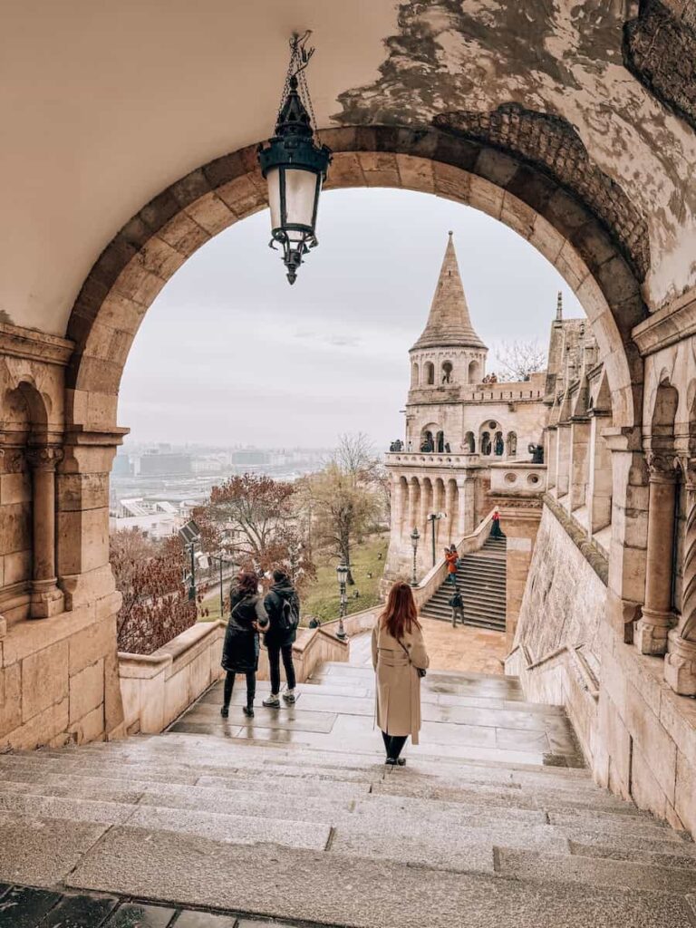 Stone archway at Fisherman’s Bastion framing visitors walking down stairs with views over Budapest on a cloudy winter day