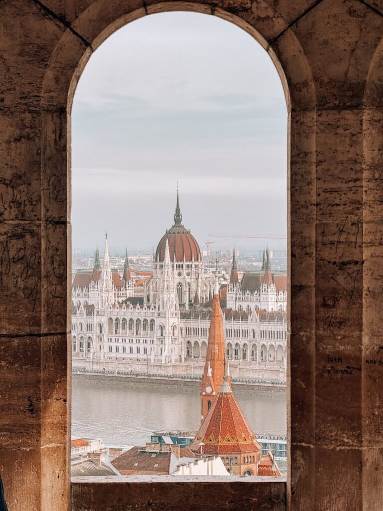 View of the Hungarian Parliament Building framed through an arched window at Fisherman’s Bastion overlooking the Danube River
