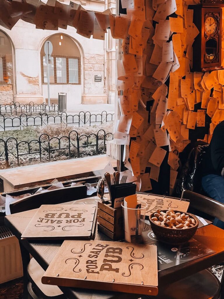 Cozy table inside the For Sale Pub in Budapest with wooden menus, a bowl of peanuts, and walls covered in handwritten notes, looking out toward a quiet street through a window.
