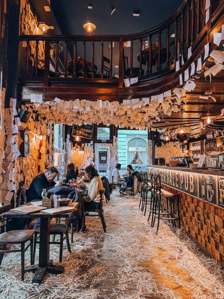 Warm rustic interior of the For Sale Pub in Budapest with straw covered floors, wooden tables and chairs, diners enjoying food and drinks, and the bar lined with notes and hanging papers under soft ambient lighting.