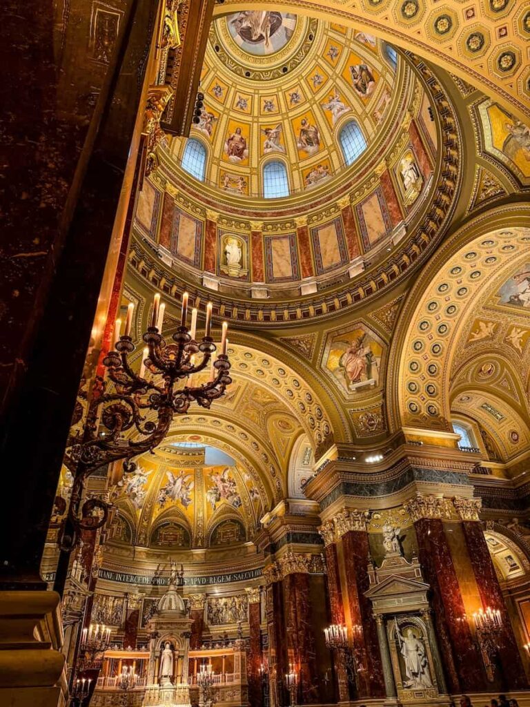 Interior of St Stephen’s Basilica in Budapest showcasing the ornate golden dome, detailed frescoes, marble columns, statues, and glowing chandeliers creating a grand and reverent atmosphere.
