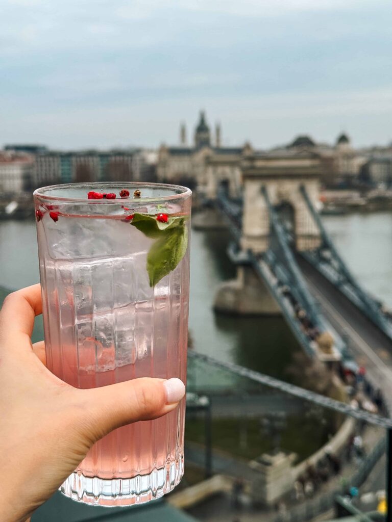 Hand holding a pink cocktail on a rooftop terrace with a view of the Chain Bridge and the Danube River in Budapest, with city buildings in the background