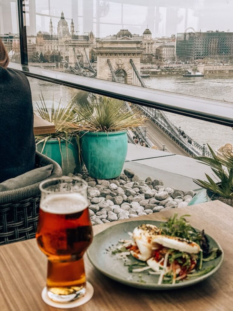 Plate of food and a glass of beer on a table inside a restaurant with large windows overlooking the Chain Bridge and the Danube River in Budapest