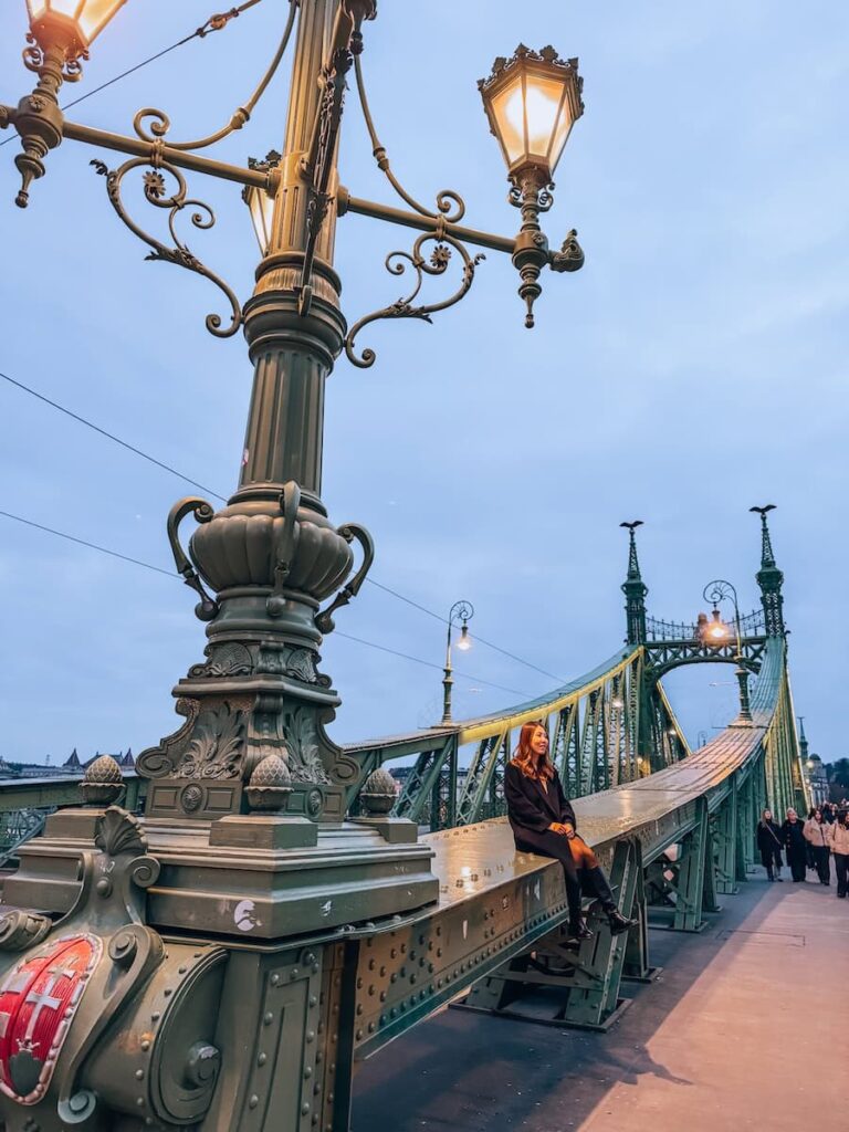 Evening view of Liberty Bridge in Budapest with decorative green metalwork and glowing street lamps, featuring a woman sitting on the bridge structure while pedestrians walk across the bridge in the distance.